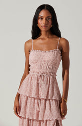 A woman is wearing a sleeveless, pink dress with a textured, ruched top and thin spaghetti straps. The dress features a waist tie and has multiple tiered, ruffled layers at the skirt.